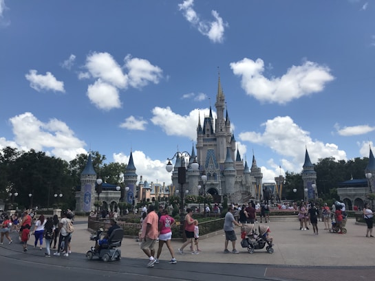 A large crowd of people is gathered in front of a fairy-tale castle with multiple spires. The sky is bright blue with scattered white clouds. Visitors are walking around, some pushing strollers, others taking photos. The scene is lively and energetic, typical of a theme park environment.
