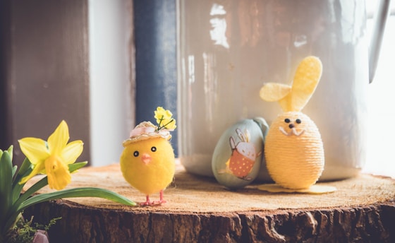 A scene with Easter-themed decorations, including a small yellow chick adorned with a floral hat and a crafted bunny figure. They are placed on a wooden surface alongside a daffodil and a decorated egg with bunny illustrations.