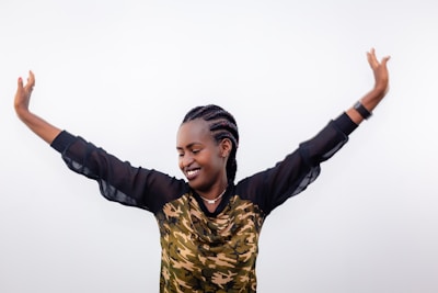 A person with braided hair wearing a camouflage-patterned top and sheer sleeves is smiling with arms raised high in a celebratory or triumphant gesture. The background is plain white, emphasizing the person's joyful expression.