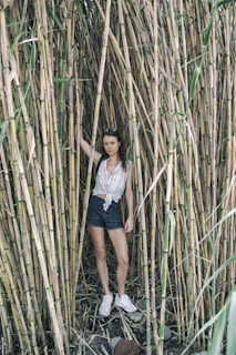 A person stands amidst tall bamboo stalks, wearing a striped, sleeveless shirt, denim shorts, and white sneakers. The person is holding one of the bamboo stalks with their right hand and appears relaxed. The ground is covered in dry leaves and rocks.