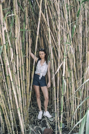 A person stands amidst tall bamboo stalks, wearing a striped, sleeveless shirt, denim shorts, and white sneakers. The person is holding one of the bamboo stalks with their right hand and appears relaxed. The ground is covered in dry leaves and rocks.