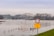 A landscape featuring a partially flooded area with a river reaching above its usual level. A bridge arches over the river in the background amidst a cloudy sky. In the foreground, a sign on a post reads a warning about the high water, with a red and white traffic barrier partially submerged.
