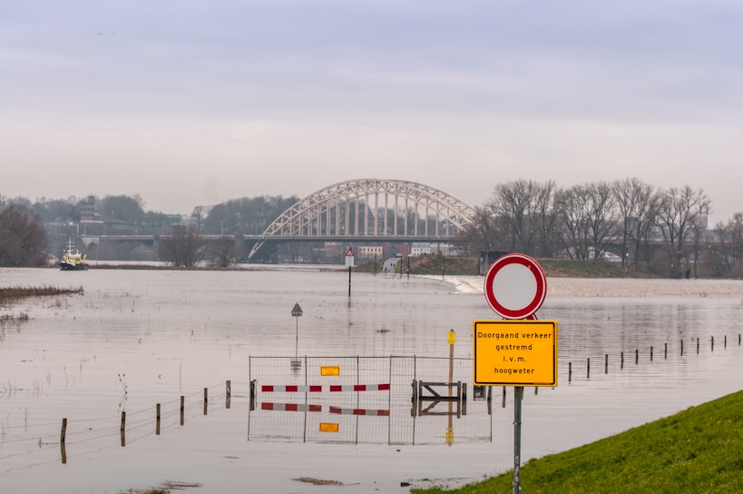 A landscape featuring a partially flooded area with a river reaching above its usual level. A bridge arches over the river in the background amidst a cloudy sky. In the foreground, a sign on a post reads a warning about the high water, with a red and white traffic barrier partially submerged.