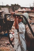 A person is standing outdoors, holding onto a metal frame. Surrounding them are piles of garbage and debris, suggesting a waste or recycling area. The person is wearing a light-colored shirt and a cap, smiling at the camera. In the background, there is another person wearing a hat, seen slightly from the side.