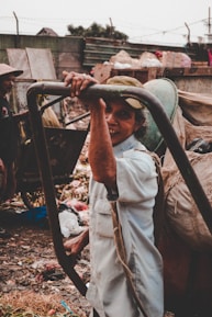 A person is standing outdoors, holding onto a metal frame. Surrounding them are piles of garbage and debris, suggesting a waste or recycling area. The person is wearing a light-colored shirt and a cap, smiling at the camera. In the background, there is another person wearing a hat, seen slightly from the side.