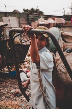 A person is standing outdoors, holding onto a metal frame. Surrounding them are piles of garbage and debris, suggesting a waste or recycling area. The person is wearing a light-colored shirt and a cap, smiling at the camera. In the background, there is another person wearing a hat, seen slightly from the side.