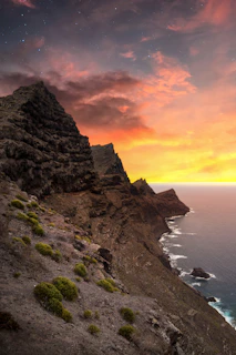 A rugged volcanic coastline at sunset in Gran Canaria, with warm orange and deep petrol hues blending into the sky.