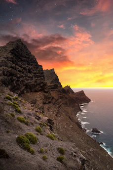 A rugged volcanic coastline at sunset in Gran Canaria, with warm orange and deep petrol hues blending into the sky.