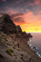 A rugged coastline with steep cliffs descending into the ocean, set against a dramatic sunset sky filled with vibrant oranges, pinks, and purples. Sparse vegetation covers the rocky terrain, adding a touch of green to the landscape. The gentle wave patterns can be observed as they meet the shore.