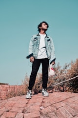 A stylish young man wearing a casual denim jacket and white sneakers, standing confidently in an urban setting.