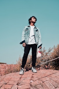 A stylish young man wearing a casual denim jacket and white sneakers, standing confidently in an urban setting.