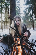 A person with long blonde hair is sitting in a snowy forest, tending to a campfire built with an arrangement of sticks and logs. The individual appears to be focused on the fire, with trees surrounding the scene.