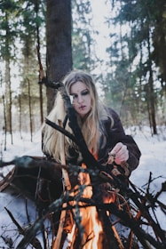 A person with long blonde hair is sitting in a snowy forest, tending to a campfire built with an arrangement of sticks and logs. The individual appears to be focused on the fire, with trees surrounding the scene.