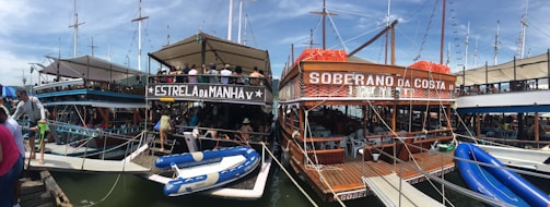 Several boats are docked at a marina, with people visible on board. The boats have wooden structures and are equipped with lifebuoys. Blue sky and masts of other boats are visible in the background. Inflatable boats are tied to the main vessels.
