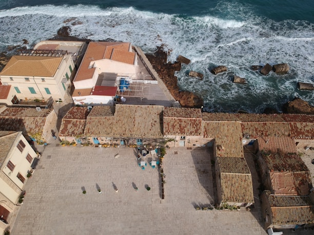 Aerial view of a coastal area with traditional buildings featuring terracotta tiled roofs. The structures are aligned alongside a paved courtyard, and in the background, waves crash against the rocky shoreline. Some outdoor furniture, including tables and chairs, is visible in the courtyard.