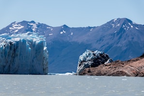 A large glacier with jagged ice formations rises from a cold, light-colored body of water. In the background, towering mountains capped with patches of snow create a dramatic contrast against the clear blue sky. The scene is serene and remote, emphasizing the vastness of nature.