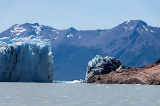 A large glacier with jagged ice formations rises from a cold, light-colored body of water. In the background, towering mountains capped with patches of snow create a dramatic contrast against the clear blue sky. The scene is serene and remote, emphasizing the vastness of nature.