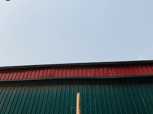 A corrugated metal roof seen from below, displaying a green panel with a red trim against a clear sky. A wooden pole is visible extending vertically up the green section.