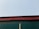 A corrugated metal roof seen from below, displaying a green panel with a red trim against a clear sky. A wooden pole is visible extending vertically up the green section.
