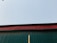 A corrugated metal roof seen from below, displaying a green panel with a red trim against a clear sky. A wooden pole is visible extending vertically up the green section.