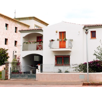 A two-story white house with a red-tiled roof and a small balcony adorned with potted plants. The house has orange-framed windows and a wrought iron railing. The ground floor features a gated entrance and plants are visible around the property. Adjacent to the house is another building with a beige facade. A lamp post and some trees or bushes are also present.
