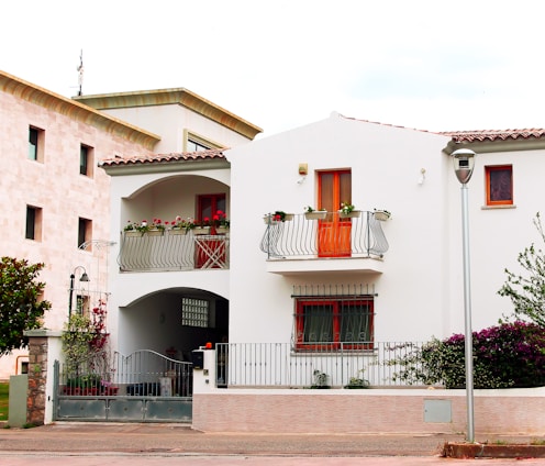 A two-story white house with a red-tiled roof and a small balcony adorned with potted plants. The house has orange-framed windows and a wrought iron railing. The ground floor features a gated entrance and plants are visible around the property. Adjacent to the house is another building with a beige facade. A lamp post and some trees or bushes are also present.