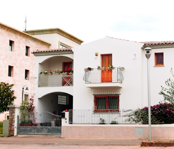A two-story white house with a red-tiled roof and a small balcony adorned with potted plants. The house has orange-framed windows and a wrought iron railing. The ground floor features a gated entrance and plants are visible around the property. Adjacent to the house is another building with a beige facade. A lamp post and some trees or bushes are also present.