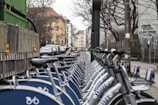 A row of parked rental bicycles lines a city street, with buildings and overcast skies in the background. Wooden fences and graffiti can be seen along the side, and a few cars are visible on the road.