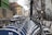 A row of parked rental bicycles lines a city street, with buildings and overcast skies in the background. Wooden fences and graffiti can be seen along the side, and a few cars are visible on the road.