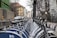 A row of parked rental bicycles lines a city street, with buildings and overcast skies in the background. Wooden fences and graffiti can be seen along the side, and a few cars are visible on the road.