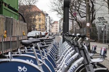 A row of parked rental bicycles lines a city street, with buildings and overcast skies in the background. Wooden fences and graffiti can be seen along the side, and a few cars are visible on the road.