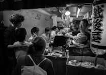 A small, cozy bar or restaurant with several people sitting at the counter, engaged in conversation. The interior features various kitchen utensils, dishes, and bottles. A man is behind the counter, appearing to clean or prepare something. The atmosphere seems lively and intimate.