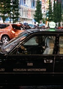 A black taxi with the signage 'KOKUSAI MOTORCARS' on its side is driving in a bustling city street at night. The taxi driver is visible through the window, and other pedestrians carrying umbrellas can be seen in the background. The street is lined with tall buildings and illuminated by bright city lights.