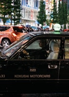A black taxi with the signage 'KOKUSAI MOTORCARS' on its side is driving in a bustling city street at night. The taxi driver is visible through the window, and other pedestrians carrying umbrellas can be seen in the background. The street is lined with tall buildings and illuminated by bright city lights.
