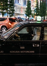 A black taxi with the signage 'KOKUSAI MOTORCARS' on its side is driving in a bustling city street at night. The taxi driver is visible through the window, and other pedestrians carrying umbrellas can be seen in the background. The street is lined with tall buildings and illuminated by bright city lights.