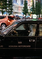 A black taxi with the signage 'KOKUSAI MOTORCARS' on its side is driving in a bustling city street at night. The taxi driver is visible through the window, and other pedestrians carrying umbrellas can be seen in the background. The street is lined with tall buildings and illuminated by bright city lights.