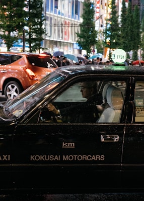 A black taxi with the signage 'KOKUSAI MOTORCARS' on its side is driving in a bustling city street at night. The taxi driver is visible through the window, and other pedestrians carrying umbrellas can be seen in the background. The street is lined with tall buildings and illuminated by bright city lights.