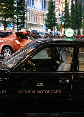 A black taxi with the signage 'KOKUSAI MOTORCARS' on its side is driving in a bustling city street at night. The taxi driver is visible through the window, and other pedestrians carrying umbrellas can be seen in the background. The street is lined with tall buildings and illuminated by bright city lights.