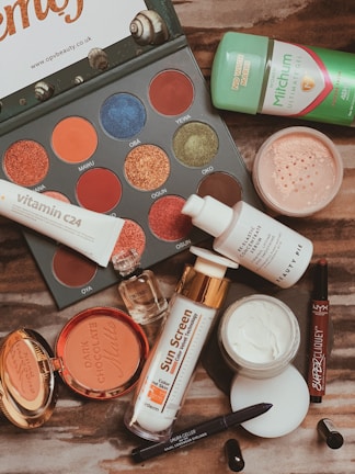 A flat lay of various beauty products including an eyeshadow palette with vibrant colors, a tube of Vitamin C serum, sunscreen, compact powder, jar of cream, lip products, a small perfume bottle, deodorant, and an eyeliner pencil. The products are arranged on a wooden surface.