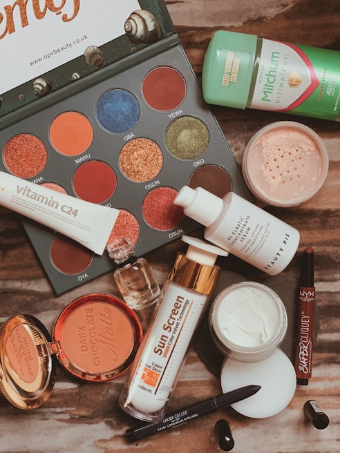 A flat lay of various beauty products including an eyeshadow palette with vibrant colors, a tube of Vitamin C serum, sunscreen, compact powder, jar of cream, lip products, a small perfume bottle, deodorant, and an eyeliner pencil. The products are arranged on a wooden surface.