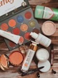 A flat lay of various beauty products including an eyeshadow palette with vibrant colors, a tube of Vitamin C serum, sunscreen, compact powder, jar of cream, lip products, a small perfume bottle, deodorant, and an eyeliner pencil. The products are arranged on a wooden surface.