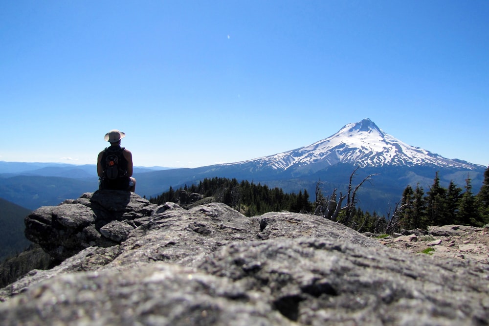 man sitting on cliff mountain viewing cone mountain covered by snow under blue sky