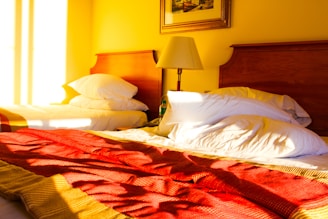 A warmly lit bedroom with two single beds. Each bed features white pillows and a red patterned bedspread. A wooden headboard is visible behind the beds, and a table lamp sits between them on a small nightstand. Above the lamp hangs a framed painting. Sunlight pours into the room from the left, casting soft, elongated shadows.
