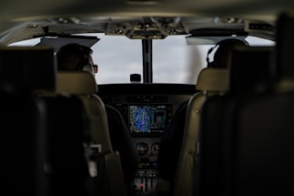 The interior of an aircraft cockpit with two pilots seated and wearing headsets. Various controls, screens, and instruments are visible in front of them, with a main display showing flight information.