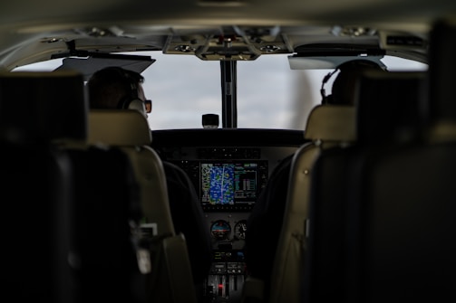 The interior of an aircraft cockpit with two pilots seated and wearing headsets. Various controls, screens, and instruments are visible in front of them, with a main display showing flight information.