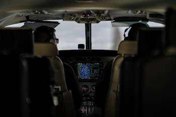The interior of an aircraft cockpit with two pilots seated and wearing headsets. Various controls, screens, and instruments are visible in front of them, with a main display showing flight information.