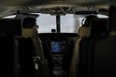 The interior of an aircraft cockpit with two pilots seated and wearing headsets. Various controls, screens, and instruments are visible in front of them, with a main display showing flight information.