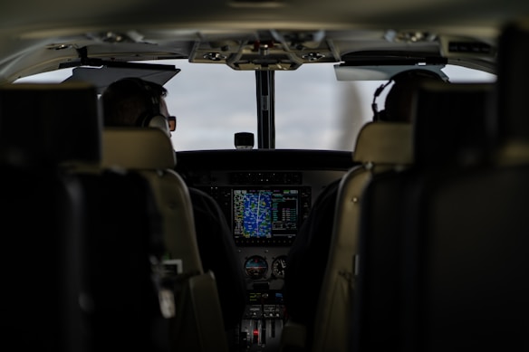 The interior of an aircraft cockpit with two pilots seated and wearing headsets. Various controls, screens, and instruments are visible in front of them, with a main display showing flight information.