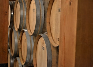 Several wooden barrels are stacked horizontally on wooden shelves. The barrels are made of a light-colored wood with metal bands securing them. Numbers are printed on the ends of the barrels, indicating identification or batch numbers. The barrels appear to be part of a larger storage facility, possibly a winery or distillery.