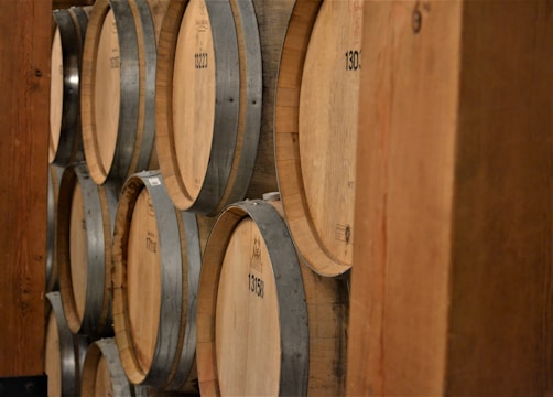 Several wooden barrels are stacked horizontally on wooden shelves. The barrels are made of a light-colored wood with metal bands securing them. Numbers are printed on the ends of the barrels, indicating identification or batch numbers. The barrels appear to be part of a larger storage facility, possibly a winery or distillery.